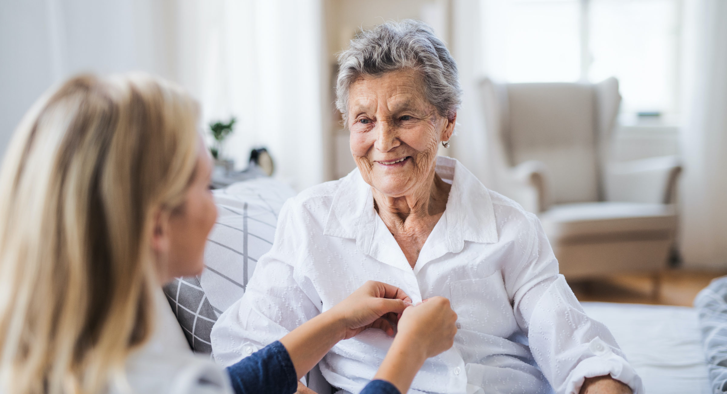 A young health visitor helping a happy sick senior woman sitting on bed at home.
